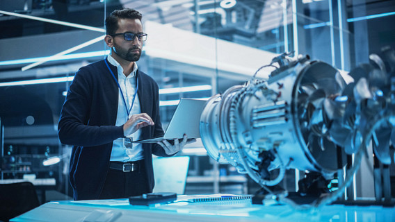 An Indian man in a modern, high-tech lab works on a laptop near a detailed turbine or jet engine model. The environment is illuminated with blue lighting, featuring glass walls, futuristic equipment, and a focused atmosphere.